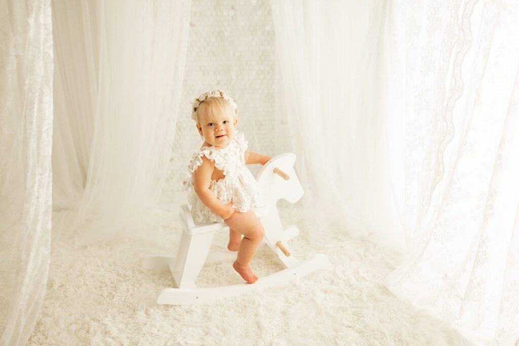 Toddler girl in a white dress and flower crown riding a rocking horse during her 1 year baby photo session in Chicago
