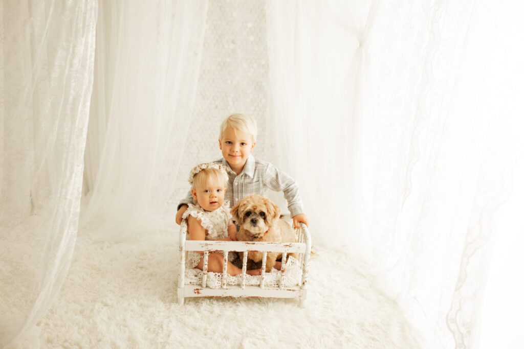 One-year-old girl and little dog in miniature crib with big brother posing behind them