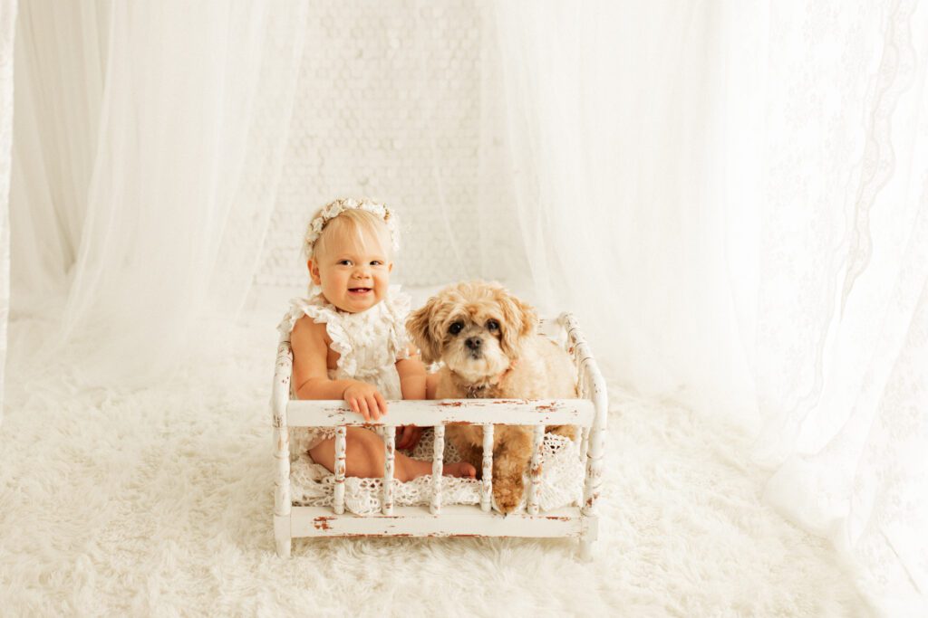 Smiling baby girl seated in antique miniature crib with her dog beside her