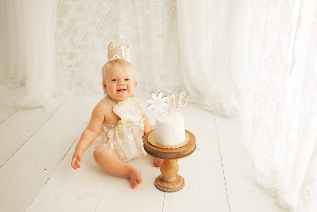 Baby girl in flowered tulle dress smiling as she sits next to her cake with a ONE cake topper