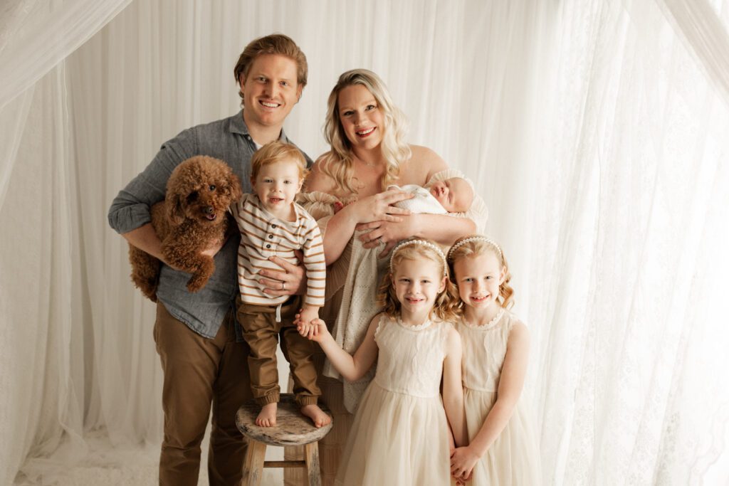 Mom and Dad pose against a lacy curtain with their four children and dog during a newborn and family session in Buffalo Grove, Illinois