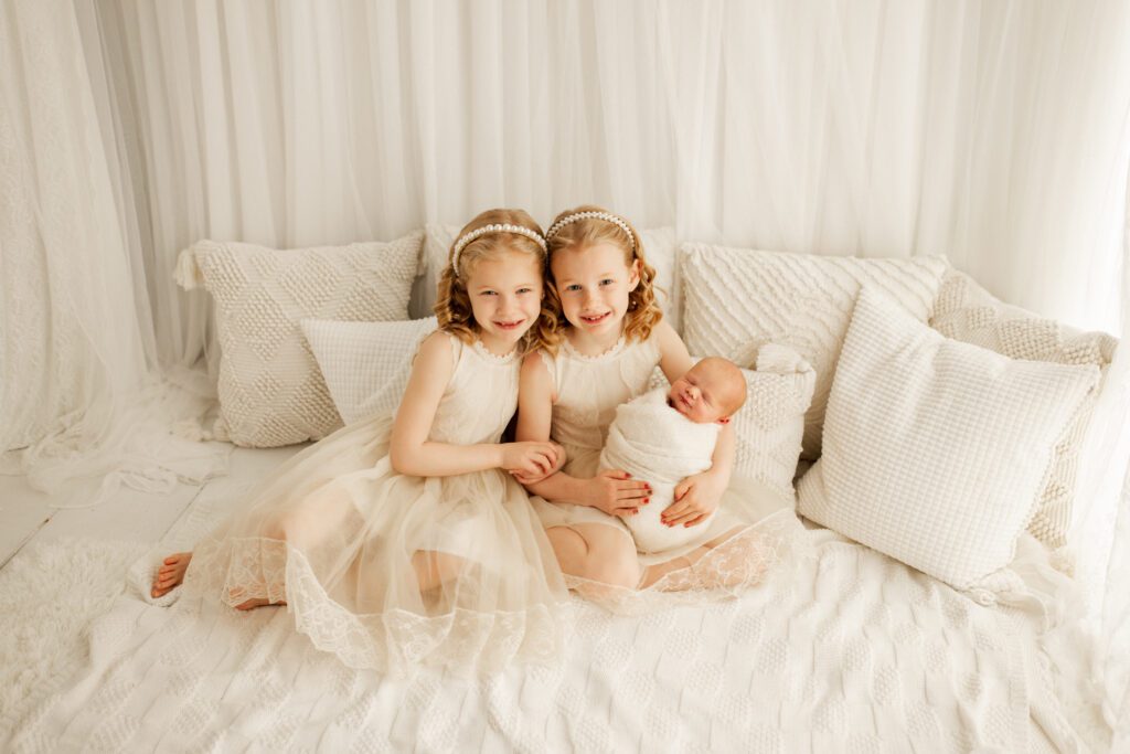 Two little girls in white dresses snuggle with their new baby brother against a backdrop of white textured pillows