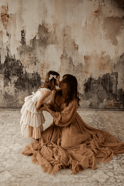 Mother in flowing beige dress and little girl in layered white dress exchange a sweet kiss during their mommy and me portraits in Chicago studio