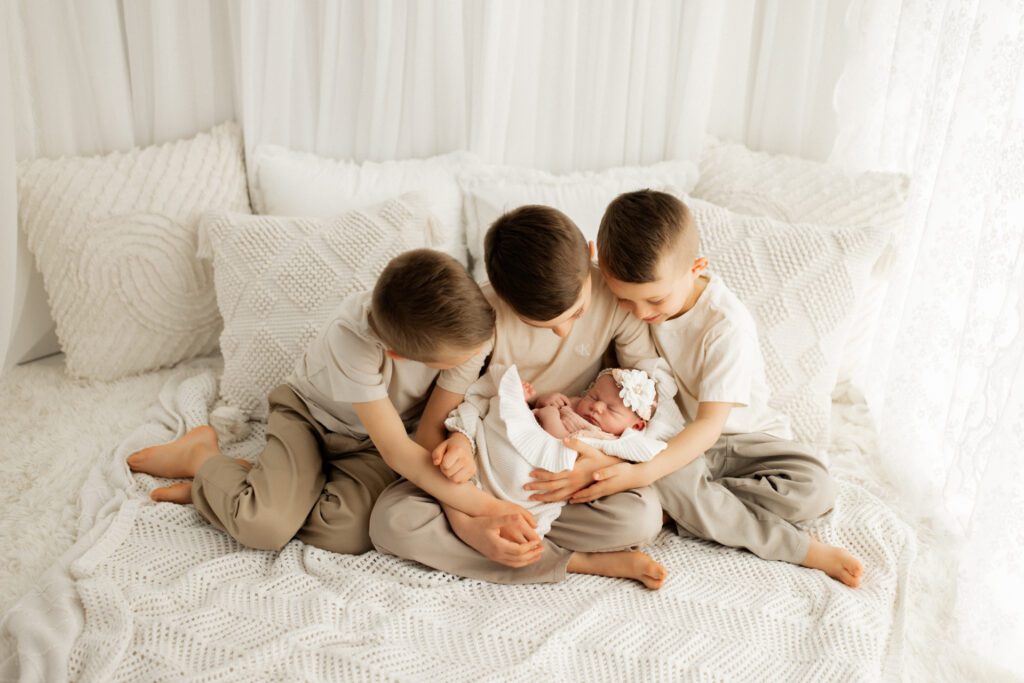 Three little boys sitting close together in a white curtained alcove snuggling their new baby sister