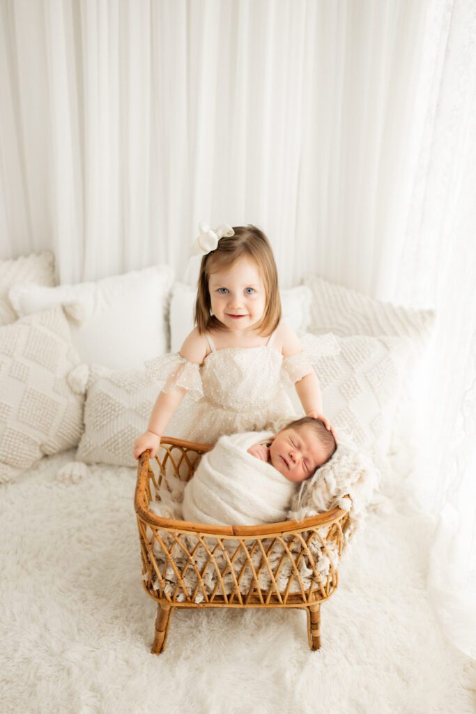 Toddler girl in white dress leaning over the bassinet where her baby brother is sleeping