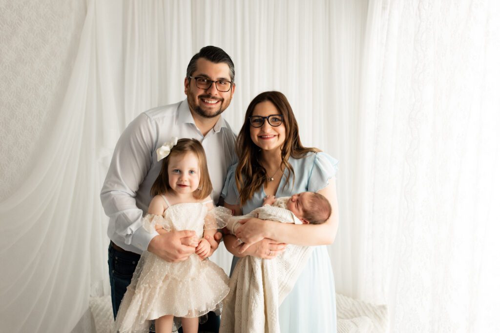 Parents with their young daughter and newborn son pose in a white curtained nook during their Vernon Hills newborn photoshoot