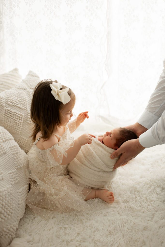 Toddler girl in white tulle dress interacting with her swaddled baby brother as dad holds him