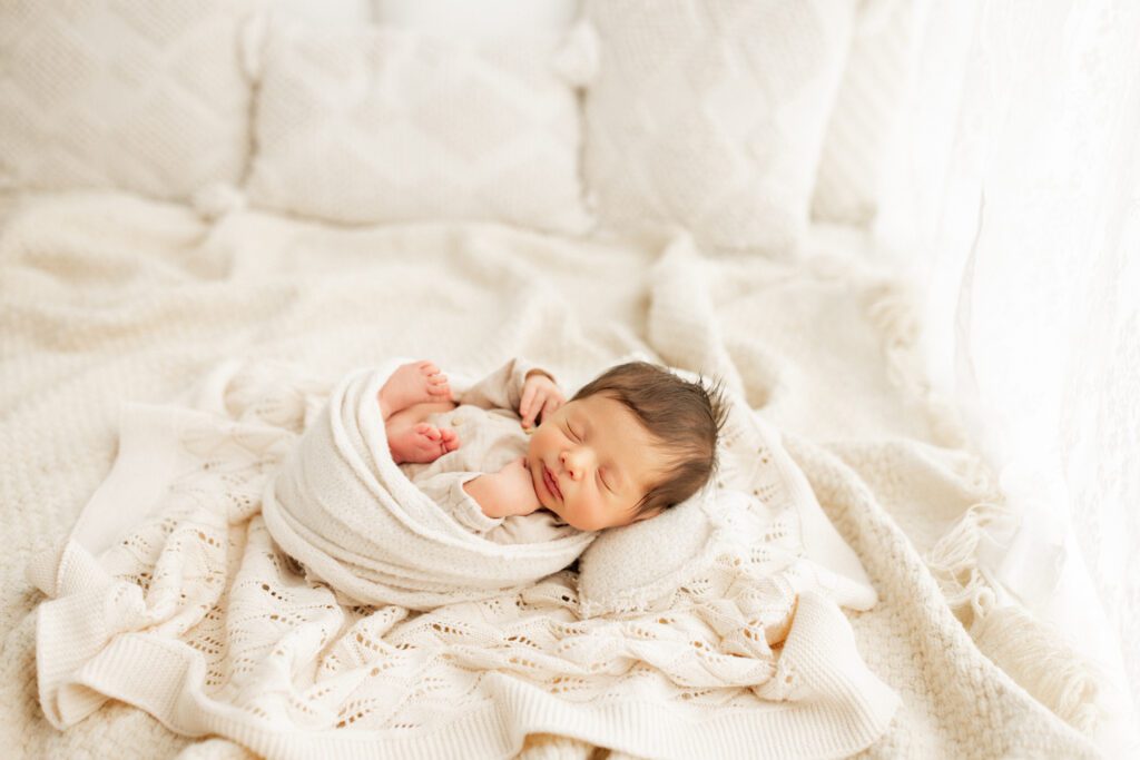 Dark-haired baby boy in cream-colored swaddle sleeping peacefully on layered blankets and pillows during his newborn photoshoot in Chicagoland studio