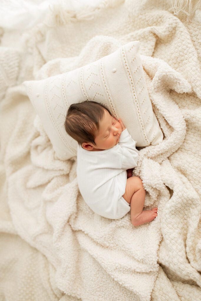 Baby boy in white onesie curled up asleep in a nest of blankets and pillows