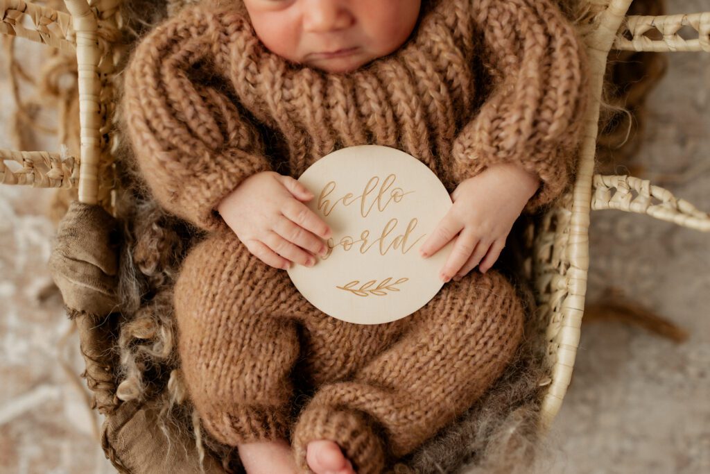 Baby boy in warm brown onesie asleep in a basket with a sign reading "Hello world" held in his hands